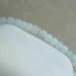 A close-up of a light blue beaded bracelet with small silver spacer beads, displayed on a white, soft textured surface against a beige background.