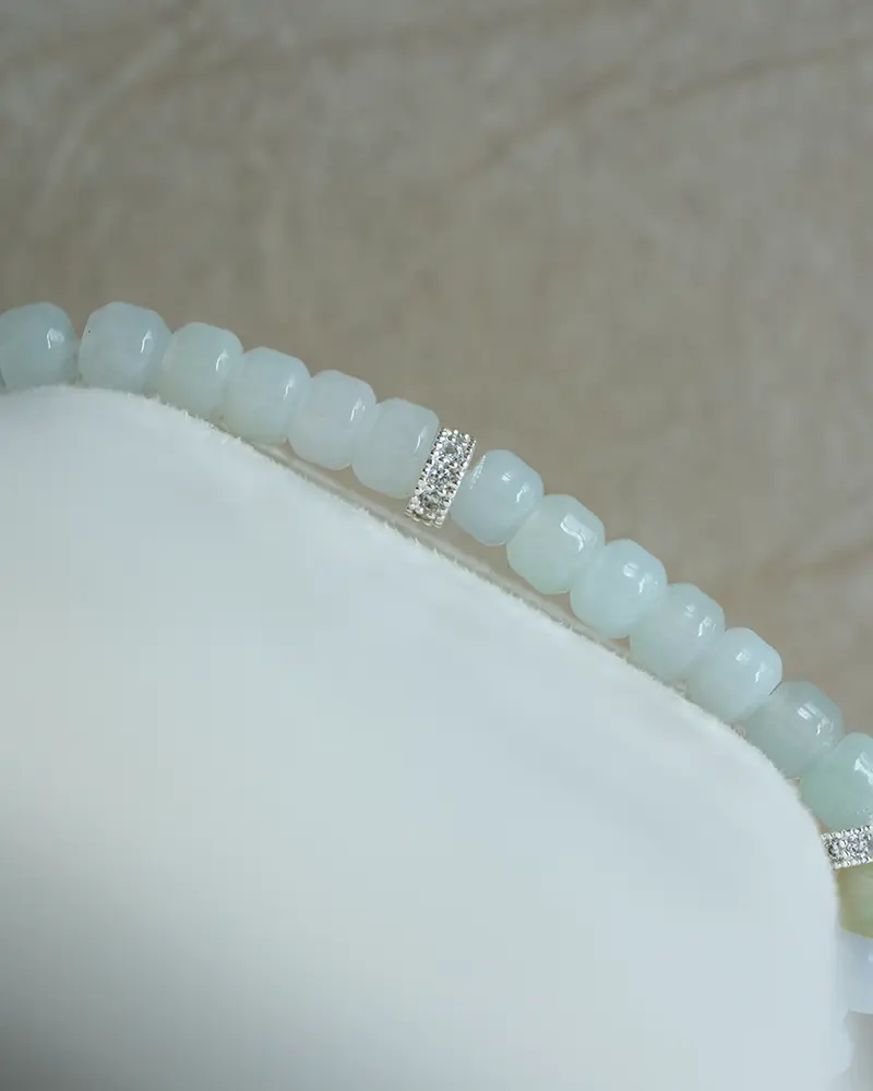 DSC_0737 拷贝 A close-up of a light blue beaded bracelet with small silver spacer beads, displayed on a white, soft textured surface against a beige background.