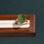 A gold ring featuring an oval green jade stone and small diamonds on the band, displayed on a wooden and cream-colored ring holder against a dark background.
