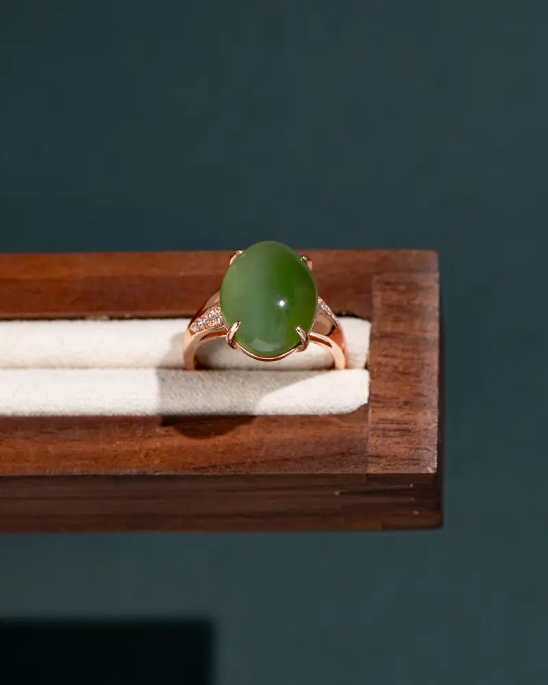 DSC_0809 拷贝 A gold ring featuring an oval green jade stone and small diamonds on the band, displayed on a wooden and cream-colored ring holder against a dark background.