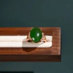 A gold ring with a large round green gemstone, possibly jade, set in the center with small clear stones on the sides, displayed on a cream-colored cushion in a wooden box.