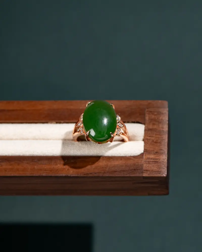 A gold ring with a large round green gemstone, possibly jade, set in the center with small clear stones on the sides, displayed on a cream-colored cushion in a wooden box.