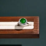A silver ring with a round green gemstone, surrounded by small clear stones, displayed on a cream-colored cushioned wooden jewelry box against a dark background.