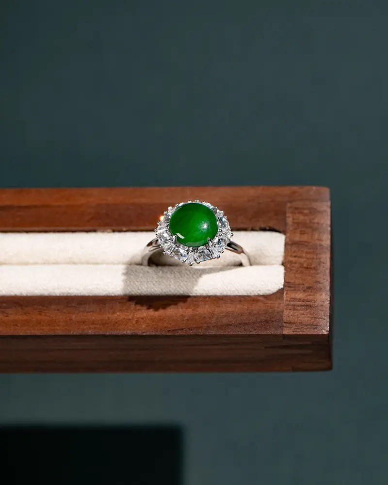 DSC_0813 拷贝 A silver ring with a round green gemstone, surrounded by small clear stones, displayed on a cream-colored cushioned wooden jewelry box against a dark background.