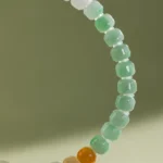 A close-up of the Brownish-Yellow, Light Green and White Jadeite Bracelet—carved beads with gold-tone accent details—set against a soft green background.