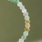 A close-up of the Brownish-Yellow, Light Green and White Jadeite Bracelet, showcasing ring-shaped beads with gold-tone accent details arranged in a gradient on an olive-green background.