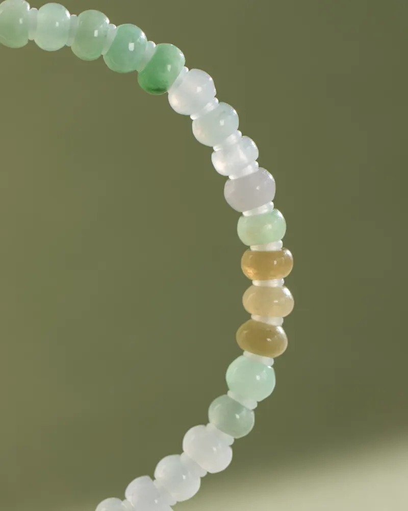 A close-up of the Brownish-Yellow, Light Green and White Jadeite Bracelet, showcasing ring-shaped beads with gold-tone accent details arranged in a gradient on an olive-green background.