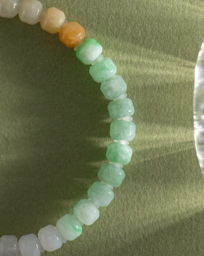 A beaded bracelet featuring brownish-yellow, light green, and white jadeite carved beads with gold-tone accent details is arranged in a partial circle on a textured green surface with sunlight casting shadows—reminiscent of the Brownish-Yellow, Light Green and White Jadeite Bracelet.