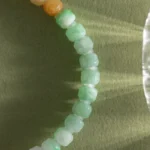 A close-up of the Brownish-Yellow, Light Green and White Jadeite Bracelet—carved beads with gold-tone accents—arranged in a curve on a textured green surface with light reflections.