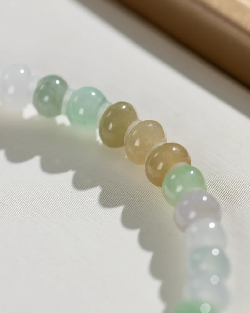 A close-up of a Brownish-Yellow, Light Green and White Jadeite Bracelet highlights ring-shaped beads in varied hues paired with gold-tone accent details, their polished surfaces casting soft shadows on a light textured background.
