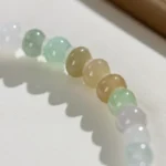 A close-up of the Brownish-Yellow, Light Green and White Jadeite Bracelet—ring-shaped beads with gold-tone accent details—shows its translucent colors casting soft shadows on a light textured surface.
