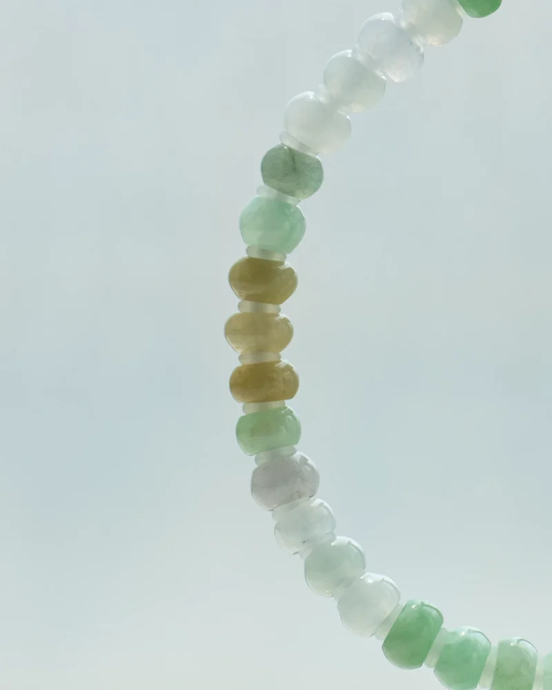 A close-up of a Brownish-Yellow, Light Green and White Jadeite Bracelet — ring-shaped beads with gold-tone accent details—against a light, blurry background.