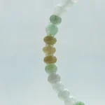 A close-up of the Brownish-Yellow, Light Green and White Jadeite Bracelet—ring-shaped beads with gold-tone accent details—arranged in a curved line against a pale, blurred background.