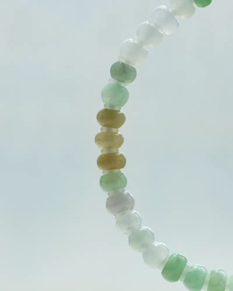 A close-up of the Brownish-Yellow, Light Green and White Jadeite Bracelet—ring-shaped beads with gold-tone accent details—arranged in a curved line against a pale, blurred background.
