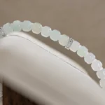 A close-up of the Light Green and White Jadeite Bracelet—featuring carved beads with silver-tone accent details—draped elegantly over a cushioned display stand.