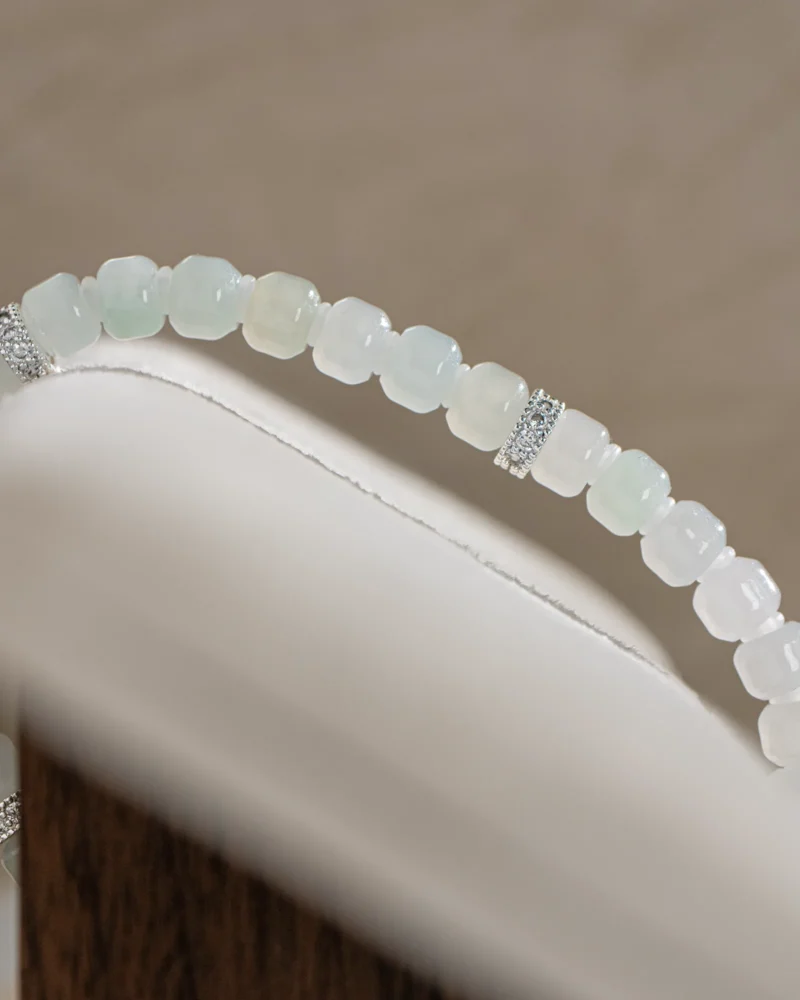 A close-up of the Light Green and White Jadeite Bracelet — Carved Beads with Silver-Tone Accent Details, showcasing translucent pale green and white beads carved into cube shapes, accented by silver-tone spacers, elegantly displayed on a white and brown cushion.