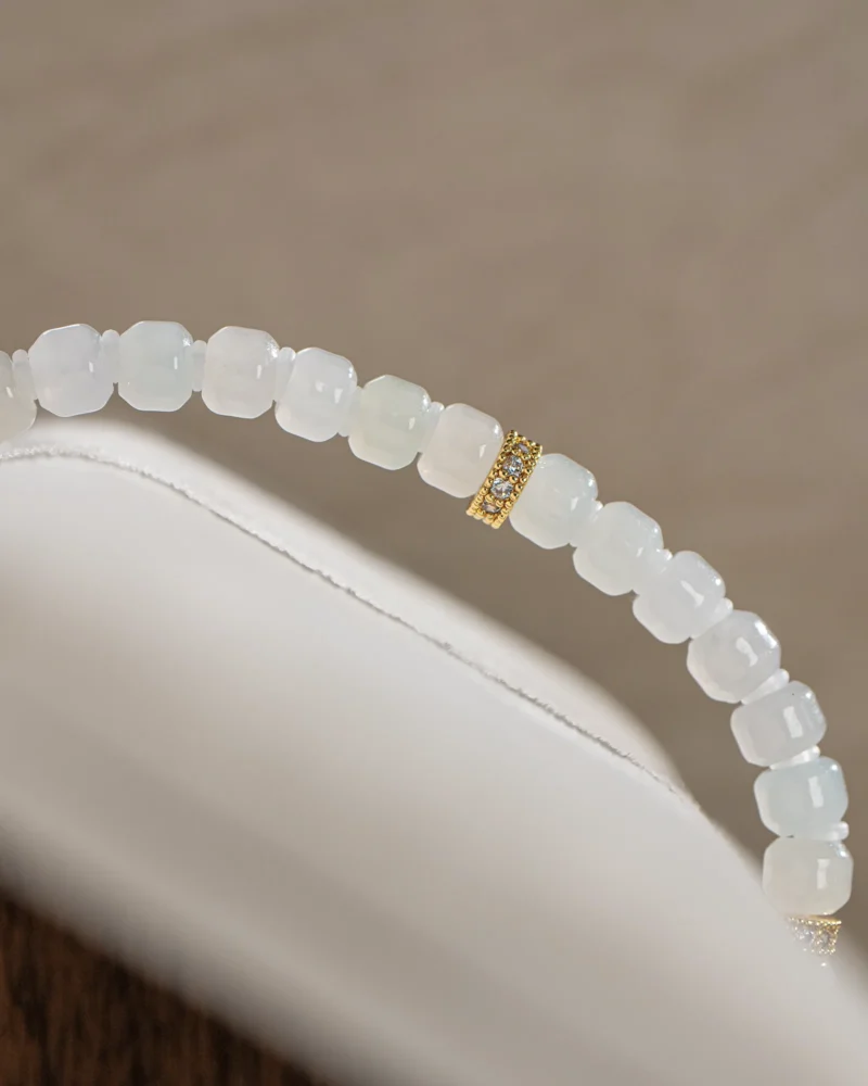A close-up of the Light Green and White Jadeite Bracelet — featuring intricately carved translucent beads and a single gold-tone accent detail, elegantly draped over a white surface.