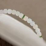 A close-up of the Light Green and White Jadeite Bracelet—carved beads and gold-tone spacer details—draped elegantly over a white curved surface.