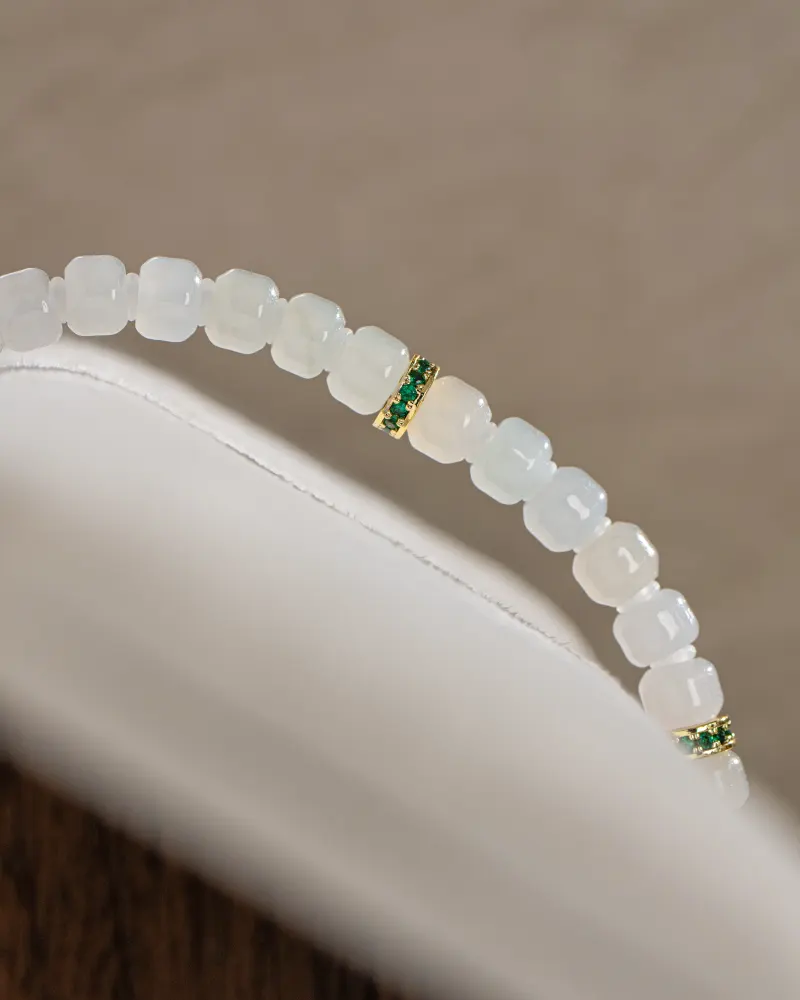 A close-up of the Light Green and White Jadeite Bracelet—carved beads and gold-tone spacer details—draped elegantly over a white curved surface.
