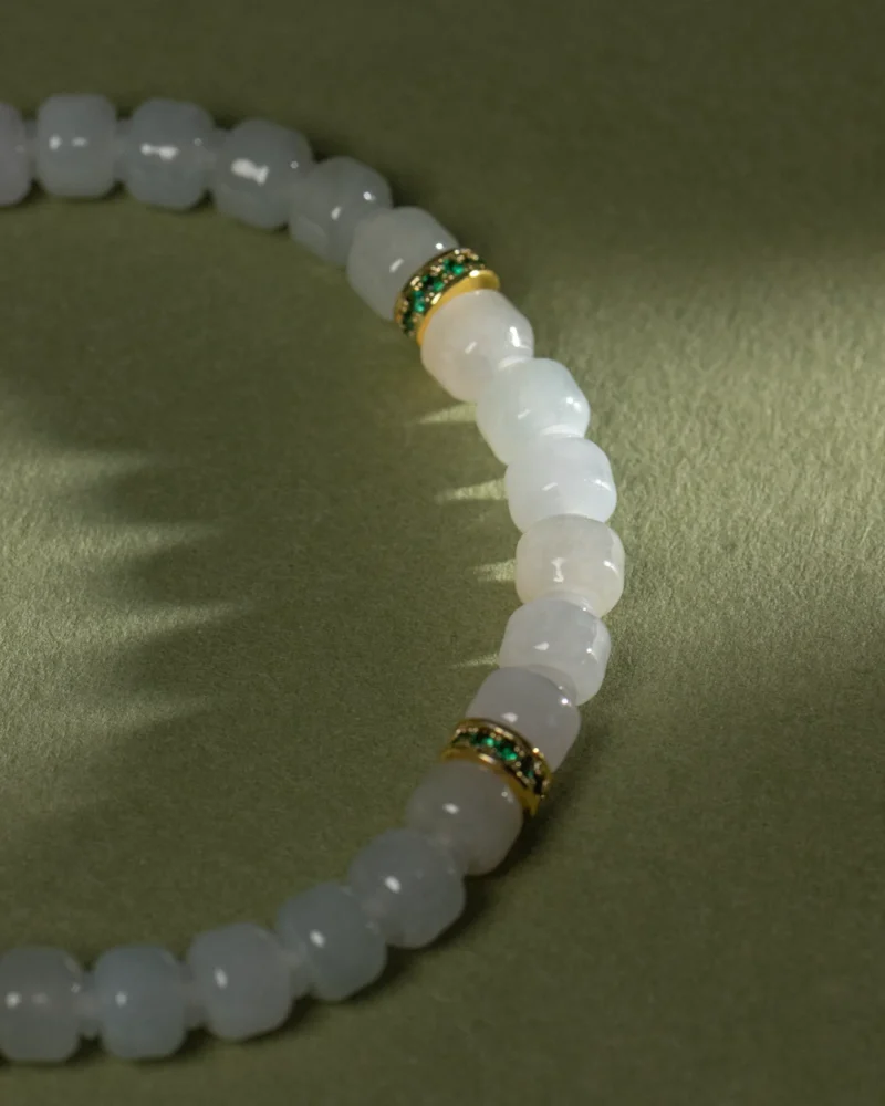 A close-up of the Light Green and White Jadeite Bracelet, showcasing intricately carved beads and gold-tone spacer details, displayed on a textured green surface.