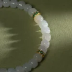 A close-up of the Light Green and White Jadeite Bracelet—carved beads with gold-tone spacer details—rests on a green surface in soft, angled light.
