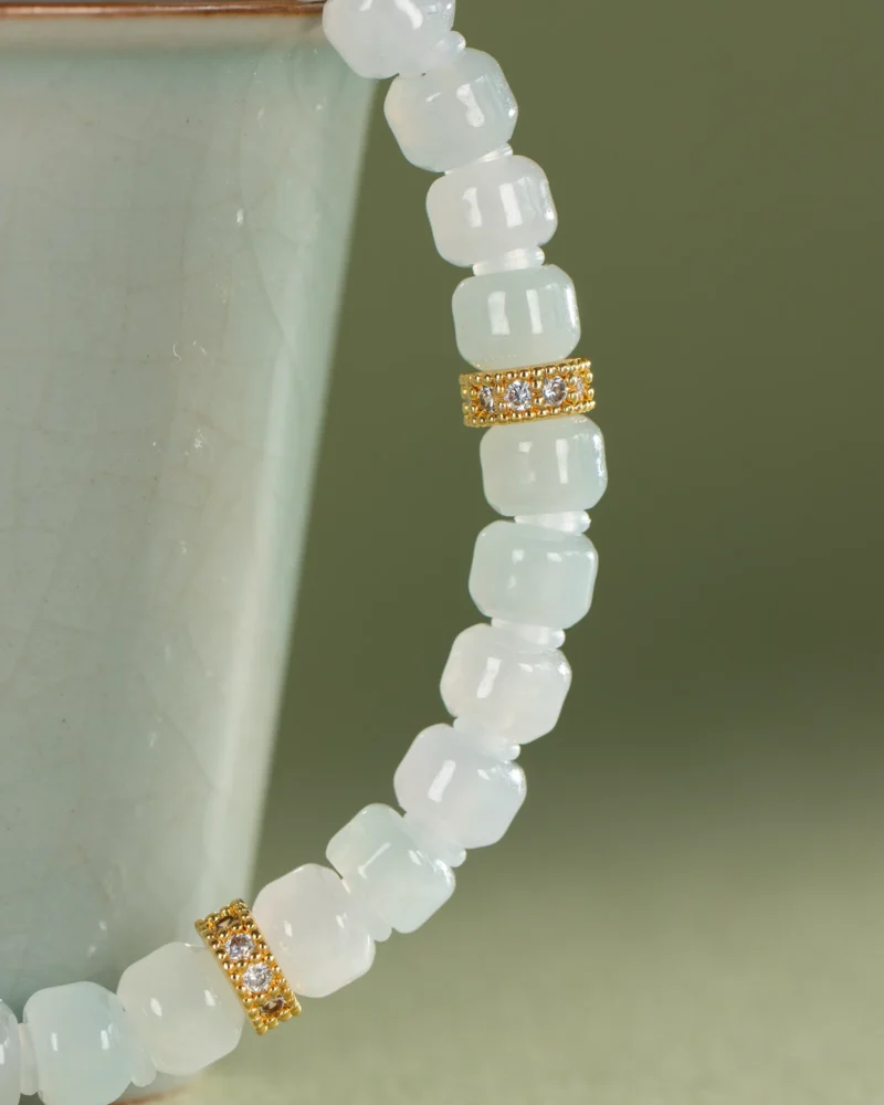 Close-up of the Light Green and White Jadeite Bracelet — Carved Beads with Gold-Tone Accent Details, showcasing its translucent beads and two gold-tone spacers adorned with delicate gems, elegantly draped on the rim of a pale green ceramic cup.