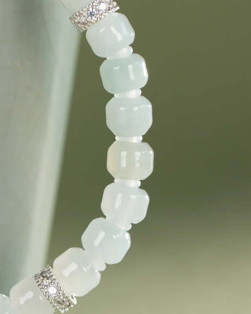A close-up of a beaded bracelet featuring light blue, faceted beads and silver spacers with small clear stones, set against a soft green background, capturing the subtle elegance of the Light Green and White Jadeite Bracelet—carved beads adorned with silver-tone accent details.
