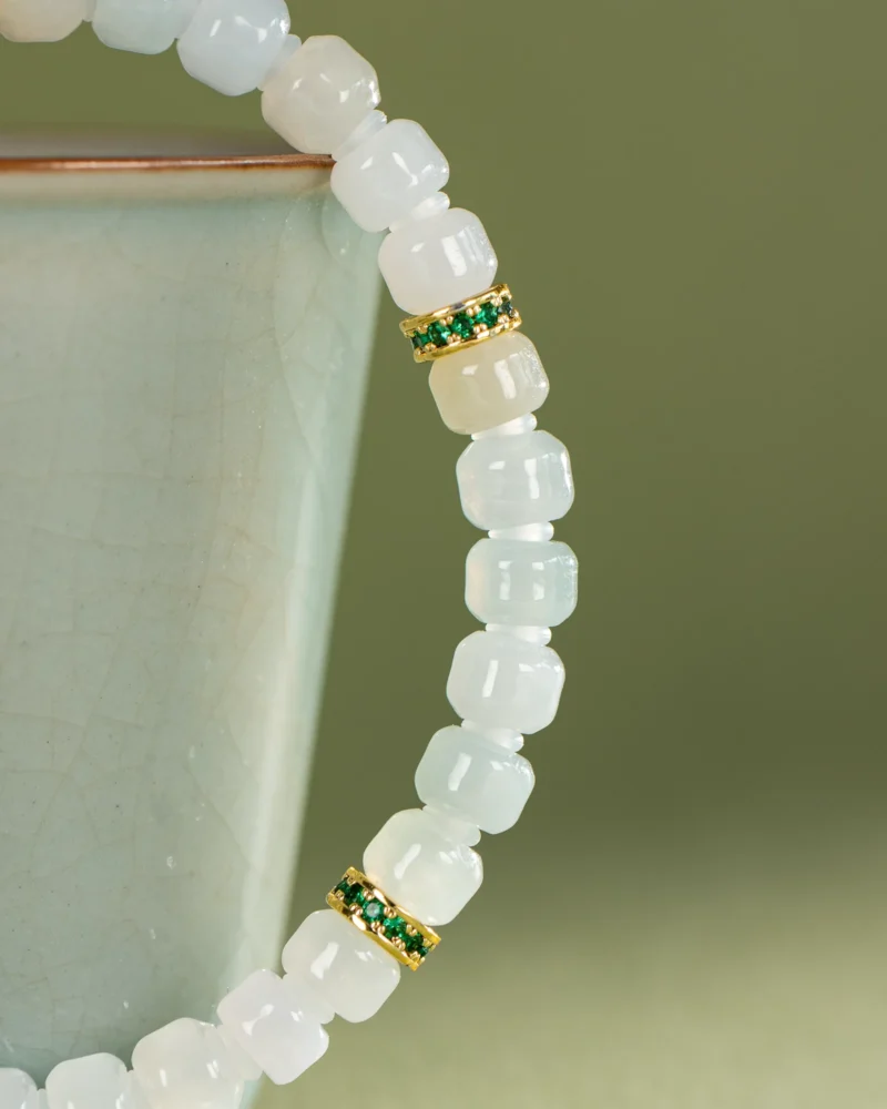 A close-up of the Light Green and White Jadeite Bracelet, featuring carved pale square beads and gold-tone spacer details, draped over the rim of a light green ceramic cup.