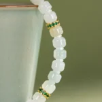 Close-up of the Light Green and White Jadeite Bracelet—carved beads with gold-tone spacers—featuring translucent white square jadeite and green-accented details, draped over a smooth, pale ceramic cup.