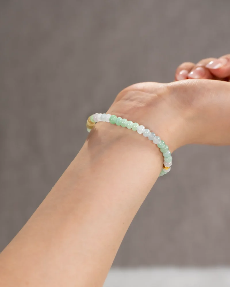 A close-up of a wrist adorned with the Brownish-Yellow, Light Green and White Jadeite Bracelet, featuring ring-shaped beads in subtle hues accented with gold-tone details, set against a softly blurred neutral background.