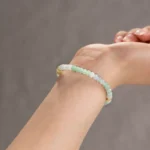 A close-up of a wrist wearing the Brownish-Yellow, Light Green, and White Jadeite Bracelet, featuring ring-shaped beads and gold-tone accent details, shown against a plain neutral background.
