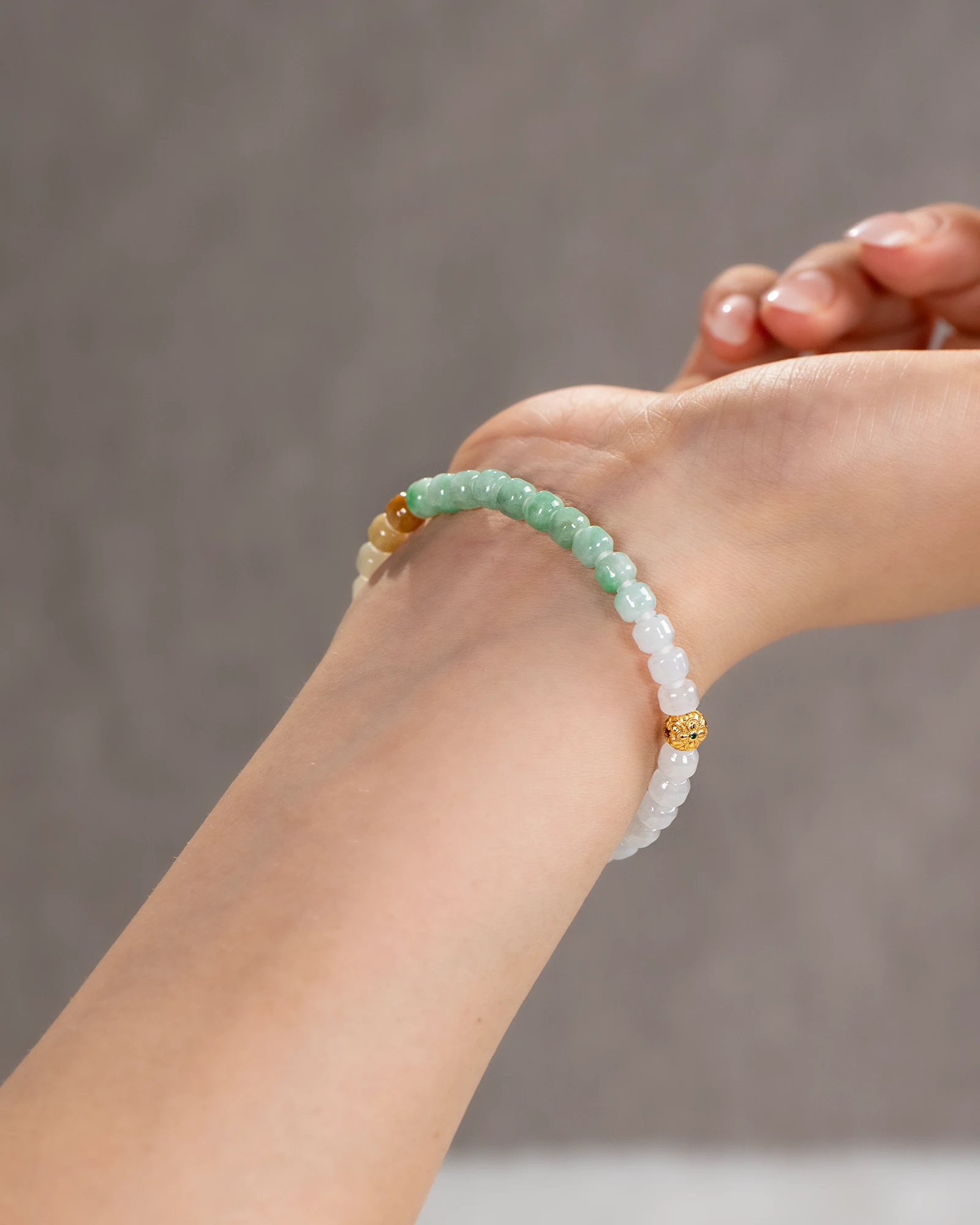 A person’s hand wearing the Brownish-Yellow, Light Green and White Jadeite Bracelet — featuring carved beads with gold-tone accent details — against a neutral background.