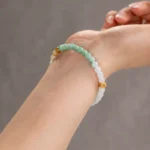 A close-up of a wrist wearing the Brownish-Yellow, Light Green and White Jadeite Bracelet—carved beads with gold-tone accent details—against a blurred gray background.