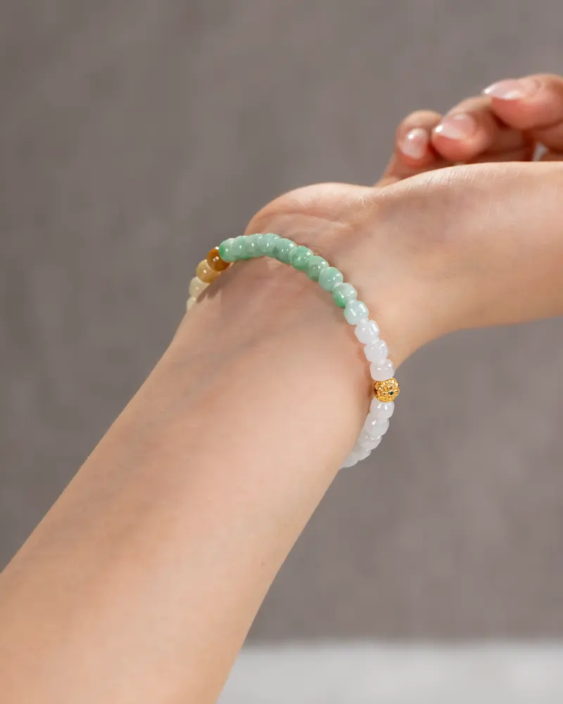 A close-up of a wrist wearing the Brownish-Yellow, Light Green and White Jadeite Bracelet—carved beads with gold-tone accent details—against a blurred gray background.