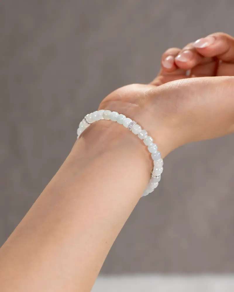 DSC_1166 拷贝 A close-up of a wrist wearing the Light Green and White Jadeite Bracelet, showcasing carved beads in pale, translucent hues with silver-tone accent details against a neutral background.