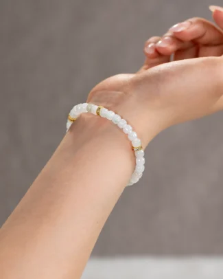 A close-up of a hand wearing the Light Green and White Jadeite Bracelet—featuring carved beads with gold-tone accent details—set against a soft, neutral background.