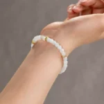 A close-up shows a person's wrist wearing the Light Green and White Jadeite Bracelet—carved beads with gold-tone accent details—set against a neutral, blurred background.
