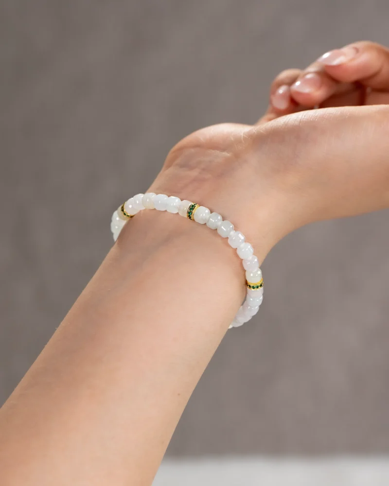 A close-up of a person’s wrist wearing the Light Green and White Jadeite Bracelet, featuring carved beads with gold-tone spacer details. The neutral background is softly out of focus.