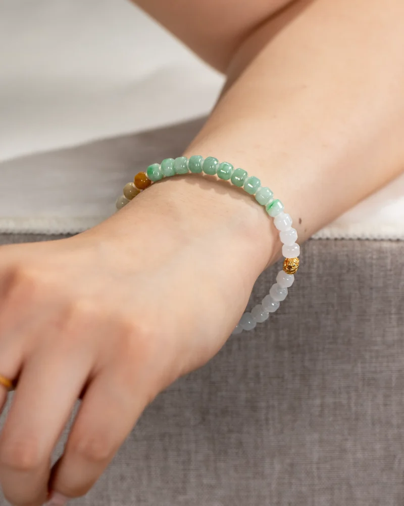 A person’s wrist wearing the Brownish-Yellow, Light Green and White Jadeite Bracelet, featuring carved beads with gold-tone accent details, with their arm resting on a gray surface.