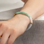A wrist wears the Brownish-Yellow, Light Green and White Jadeite Bracelet—carved beads with gold-tone accents—resting on a light grey fabric surface.