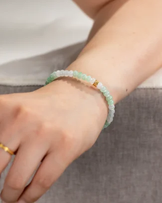 A hand wearing the Brownish-Yellow, Light Green and White Jadeite Bracelet featuring ring-shaped beads and gold-tone accent details rests on a gray surface. The person also wears a gold ring on their index finger, which complements the subtle elegance of the jadeite bracelet.