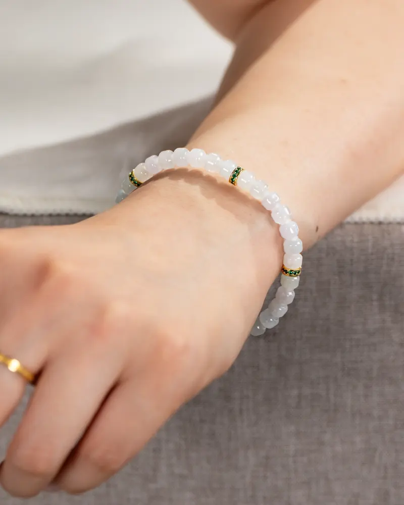 A close-up of a wrist wearing the Light Green and White Jadeite Bracelet—carved beads with gold-tone spacer details. The person also has a gold ring on their finger.