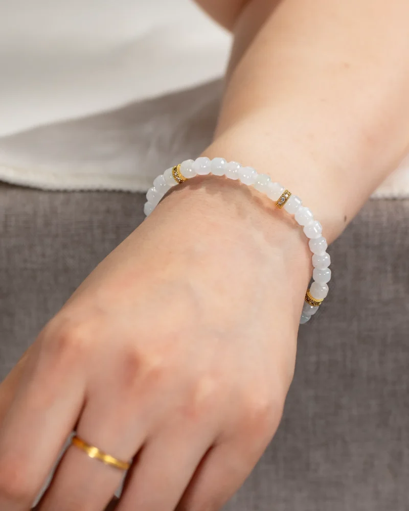 Wearing the Light Green and White Jadeite Bracelet — Carved Beads with Gold-Tone Accent Details on their wrist and a simple gold ring on their finger, the person is set against a gray surface while wearing a white top.