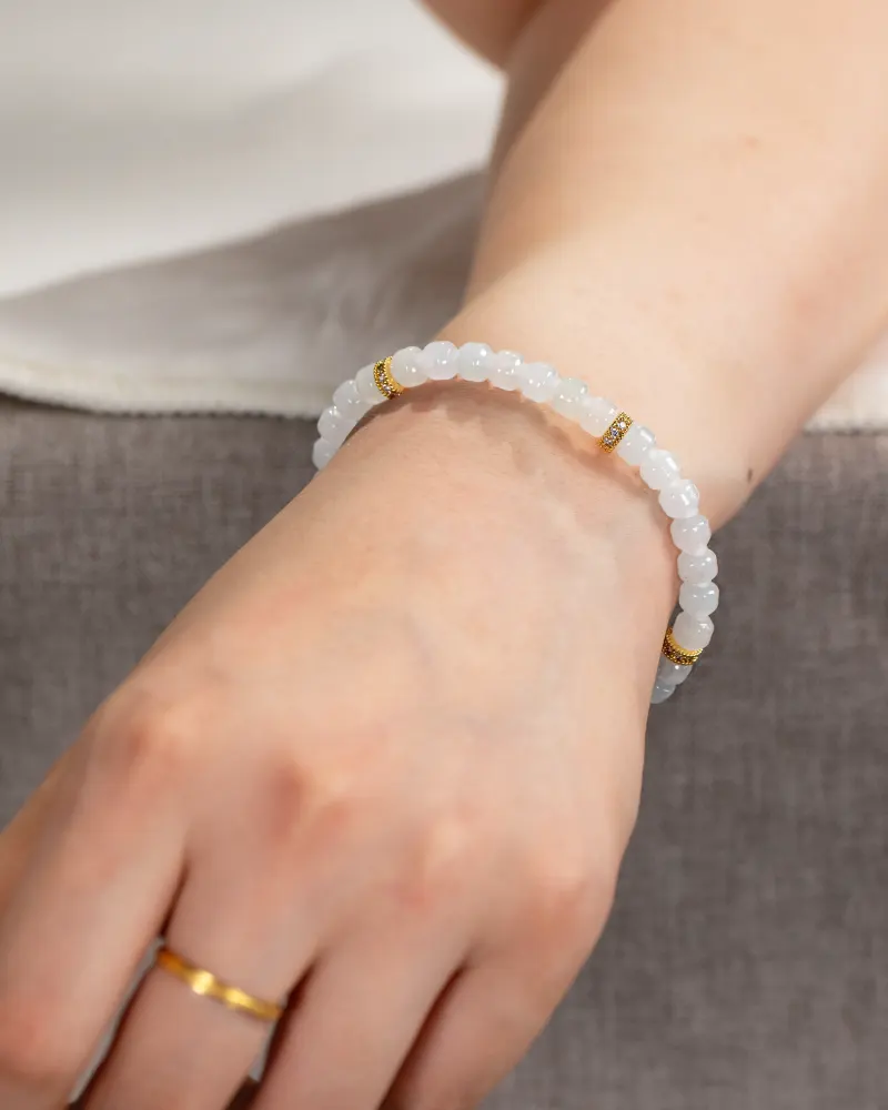 DSC_1177 拷贝 A hand wearing a gold ring and the Light Green and White Jadeite Bracelet—featuring carved beads with gold-tone accents—rests on gray fabric. The person is also wearing a white top.