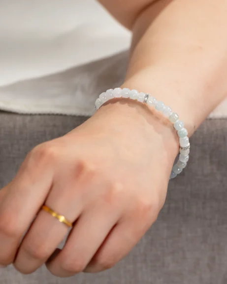 A hand resting on a gray surface is adorned with a light blue beaded bracelet featuring silver accents, the Light Green and White Jadeite Bracelet—carved beads with silver-tone accent details—and a simple gold ring on the ring finger.