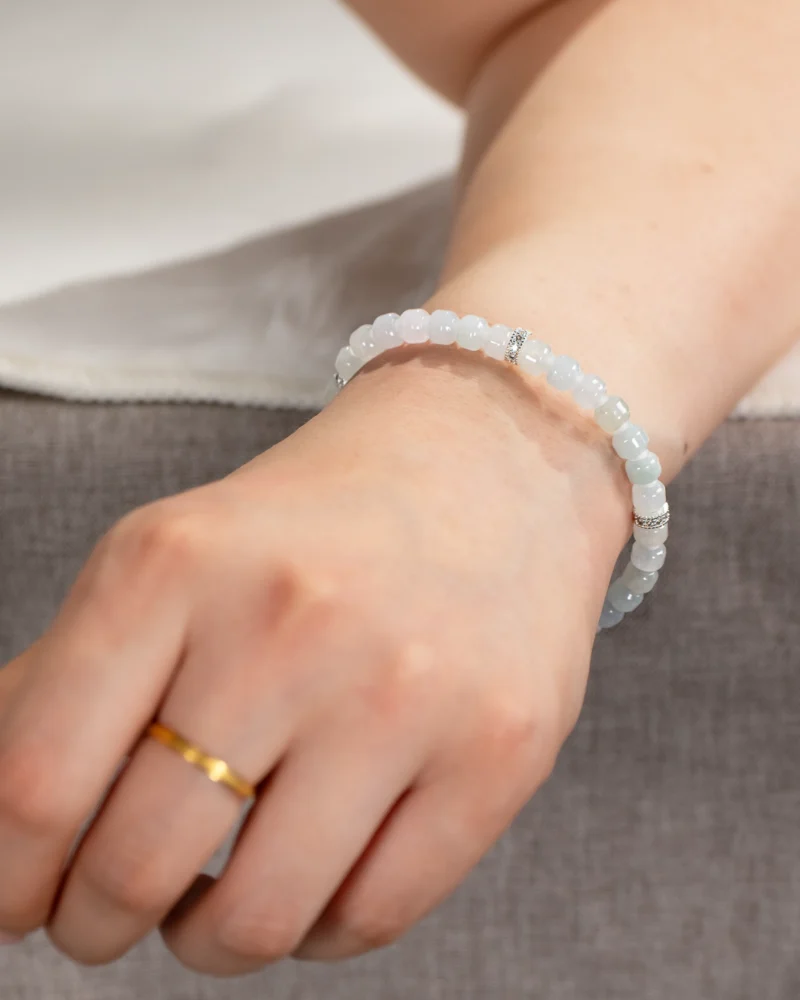 A hand resting on a gray surface is adorned with a light blue beaded bracelet featuring silver accents, the Light Green and White Jadeite Bracelet—carved beads with silver-tone accent details—and a simple gold ring on the ring finger.
