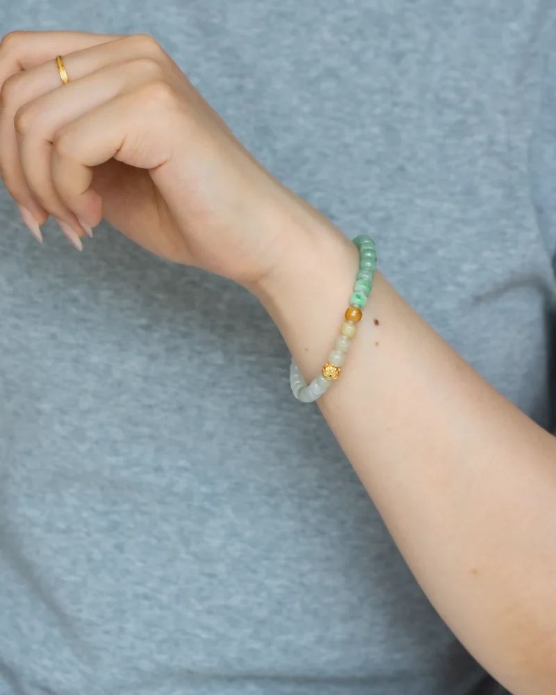 A hand wearing the Brownish-Yellow, Light Green and White Jadeite Bracelet — featuring carved beads and gold-tone accent details — along with a simple gold ring on the pinky finger. The person is dressed in a gray shirt.