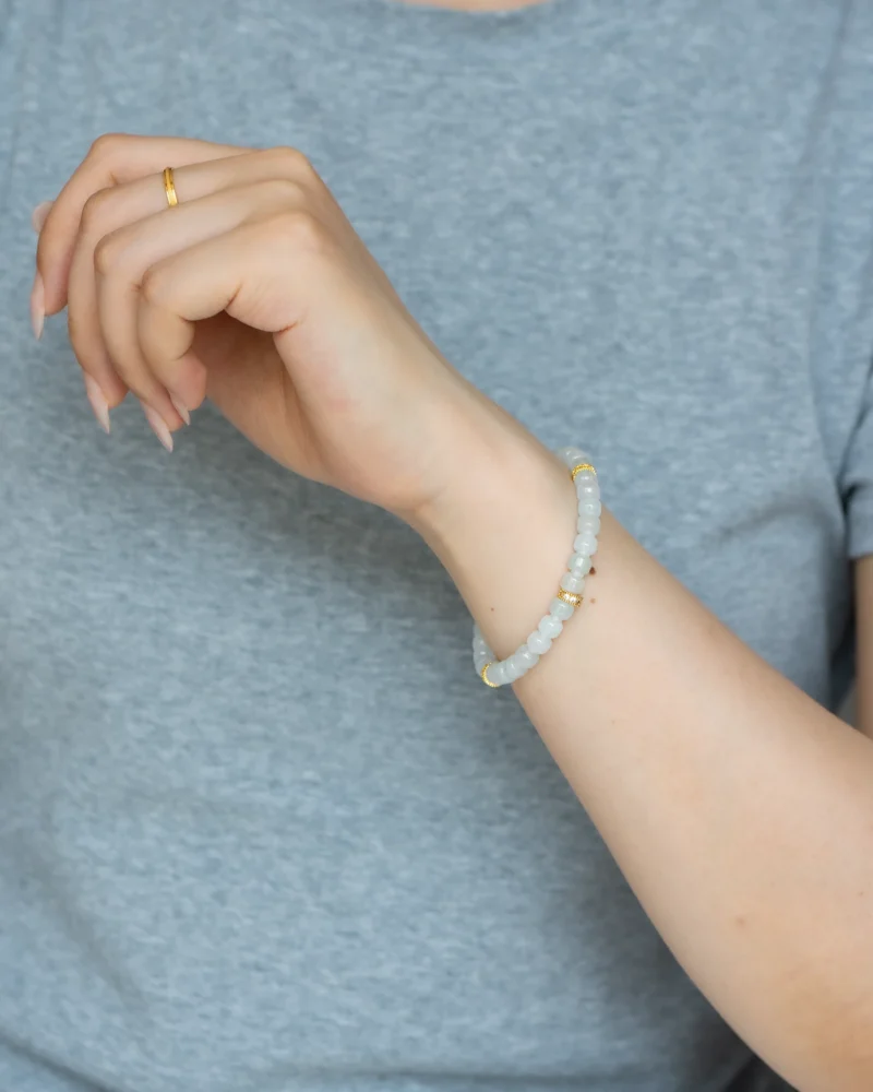 A person wearing a light gray shirt displays their left hand adorned with a simple gold ring and the Light Green and White Jadeite Bracelet — featuring carved beads with gold-tone accent details—on their wrist.