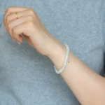 A person in a light grey top showcases their left hand, highlighting a gold ring and a Light Green and White Jadeite Bracelet—featuring carved beads with silver-tone accent details—on the wrist.