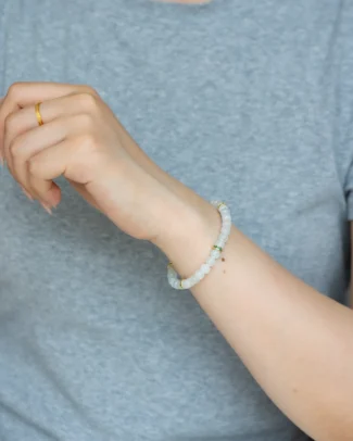 A person wearing a gray shirt has a gold ring on their finger and a Light Green and White Jadeite Bracelet—featuring carved beads with gold-tone spacer details—on their wrist.